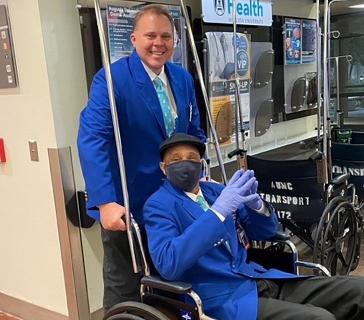Man in blue coat pushing another man in blue coat in wheelchair
