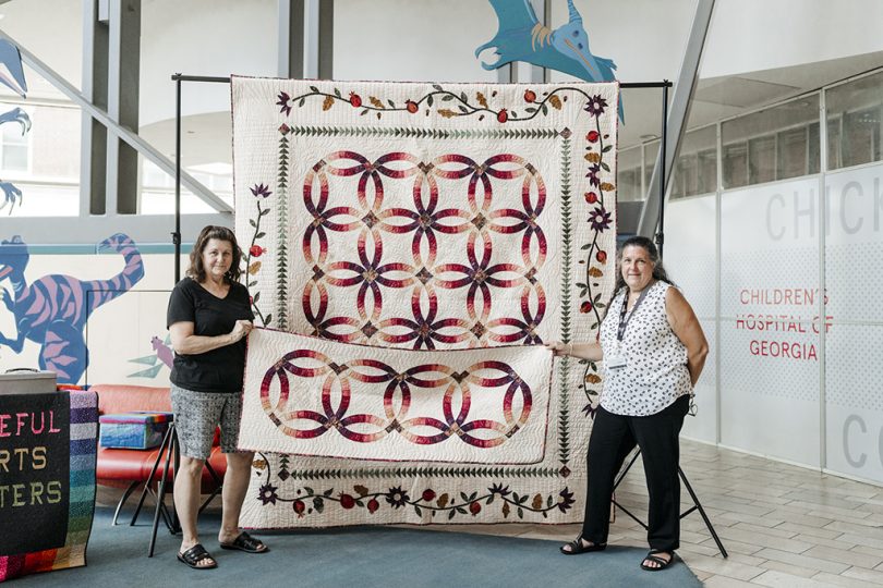 Two women stand in front of a quilt
