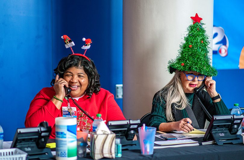 Woman in red with antlers and woman with Christmas tree hat talk on phones