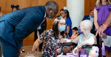 Man in suit and woman in mask talk to cancer patient in wheelchair